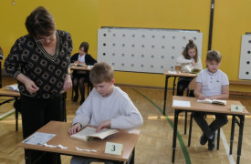 Dzieci siedzą w ławkach. Przy jednej z nich stoi kobieta 