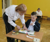 Dzieci siedzą w ławkach. Przy jednej z nich stoi kobieta