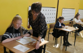 Dzieci siedzą w ławkach. Przy jednej z dziewczynek stoi kobieta