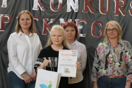 Dziewczynka prezentuje zdobyty dyplom. Za dziewczynką znajdują się trzy kobiety