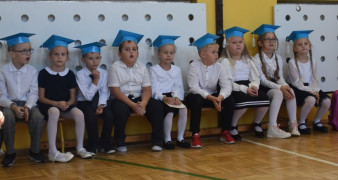 Grupa dzieci w niebieskich biretach siedzi na ławce 