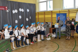 Grupa dzieci w niebieskich biretach na głowach. Widoczne dwie dorosłe osoby stojące obok dzieci
