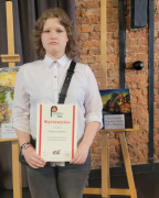Dziewczynka trzyma w ręku dyplom. Za nią znajduje się praca plastyczna przygotowana na konkurs 