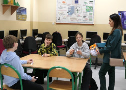 Uczniowie siedzą w szkolnych ławkach. Obok nich stoi kobieta 