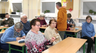 Seniorzy siedzą w szkolnych ławkach. Mężczyzna chodzi między ławkami. Trwa szkolenie dla seniorów