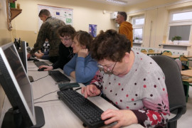 Seniorzy siedzą na krzesłach. Przed nimi znajdują się monitory. Trwa szkolenie w ramach projektu Szkoła Międzypokoleniowa 