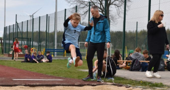 Chłopiec wykonuje skok w dal. Obserwuje go mężczyzna w sportowym dresie 