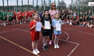 Trzy dziewczynki w strojach sportowych stoją w płycie boiska. Dziewczynki trzymają w dłoniach dyplomy. Za nimi stoi kobieta 