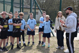 Chłopcy w sportowych strojach stoją na boisku. Obok nich znajduje się mężczyzna i kobieta. Mężczyzna tłumaczy chłopcom zasady rywalizacji sportowej 