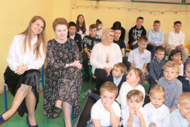 Grupa uczniów siedząca na ławeczkach. Obok na krzesłach siedzą dwie nauczycielki 