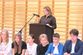 Uczniowie siedzą na ławce. Tuż za nimi stoi kobieta i przemawia do mikrofonu