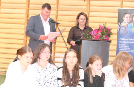 Uczniowie siedzą na ławce. Za nimi stoi kobieta i mężczyzna. Mężczyzna trzyma w ręku dokument