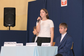 Dziewczyna i dwaj chłopcy. Chłopcy siedzą przy stoliku. Dziewczyna stoi trzymając w ręku mikrofon 