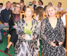 Grupa osób. Na pierwszym planie dwie kobiety siedząca na krzesłach obok siebie. Jedna z nich trzyma wrzos