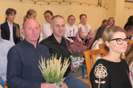 Grupa osób. Na pierwszym planie kobieta w okularach i dwa mężczyźni. Obaj trzymają wrzosy 
