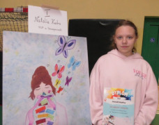 Dziewczynka stoi obok pracy konkursowej. W dłoniach trzyma dyplom 