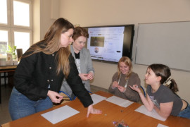 Dwie kobiety stoją. Kobieta i dziewczynka siedzą przy stole. Trwa rozmowa