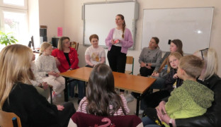 Grupa osób: dorośli i dzieci siedzą przy stole. Jedna z kobiet stoi i przemawia 