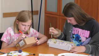 Dwie dziewczynki siedzą przy stole. Obie robią ozdobne pudełka 