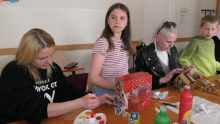 Dorośli i dzieci siedzą przy stole. Wszyscy robią ozdobne pudełka 