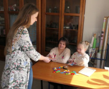 Dwie kobiety i dziewczynka. Jedn z kobiet siedzi w ławce z dziewczynką. Druga kobieta stoi obok ławki 