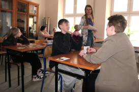 Dorośli i dzieci siedzą w szkolnych ławkach. Jedna z kobiet stoi. Trwają warsztaty 