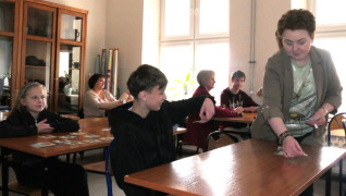 Grupa osób podczas warsztatów edukacyjnych. Większość z nich siedzi w ławkach. Jedna kobieta stoi przy ławce 