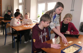 Dwie kobiety i chłopiec. Kobieta i chłopiec siedzą w szkolnej ławce. Między nimi stoi kobieta. Trwają warsztaty edukacyjne 