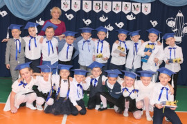 Grupa osób: uczniowie w granatowych biretach na głowie. Wśród nich stoi nauczycielka w bordowej sukience 