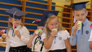 Dwie dziewczynki i chłopiec stoją obok siebie. Wszyscy w dłoniach trzymają kubeczki, a na głowach mają granatowe birety  