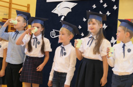 Grupa dzieci w granatowych biretach na głowie stoi obok siebie. Maluchy na głowach mają granatowe birety, a w ręku trzymają kubki