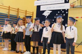 Grupa dzieci w granatowych biretach na głowie stoi obok siebie