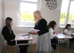 Uczniowie siedzą w szkolnych ławkach. Kobieta rozdaje im arkusze egzaminacyjne 