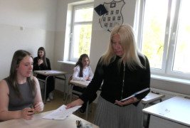 Uczniowie siedzą w szkolnych ławkach. Kobieta rozdaje im arkusze egzaminacyjne 