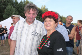 Kobieta w czerwonym wianku na głowie. Obok kobiety stoi mężczyzna w koszuli w paski 