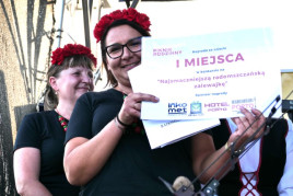 Dwie kobiety w czerwonych wiankach na głowie. Jedna z nich trzyma w ręku białą tabliczkę z czerwonymi napisami 